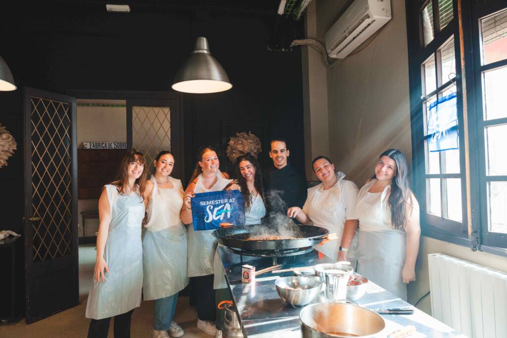 Seven people in white plastic aprons stand around a metal counter covered in cooking utensils and a large, steaming pot of food. Two people hold up a small blue flag that says “Semester at Sea.”