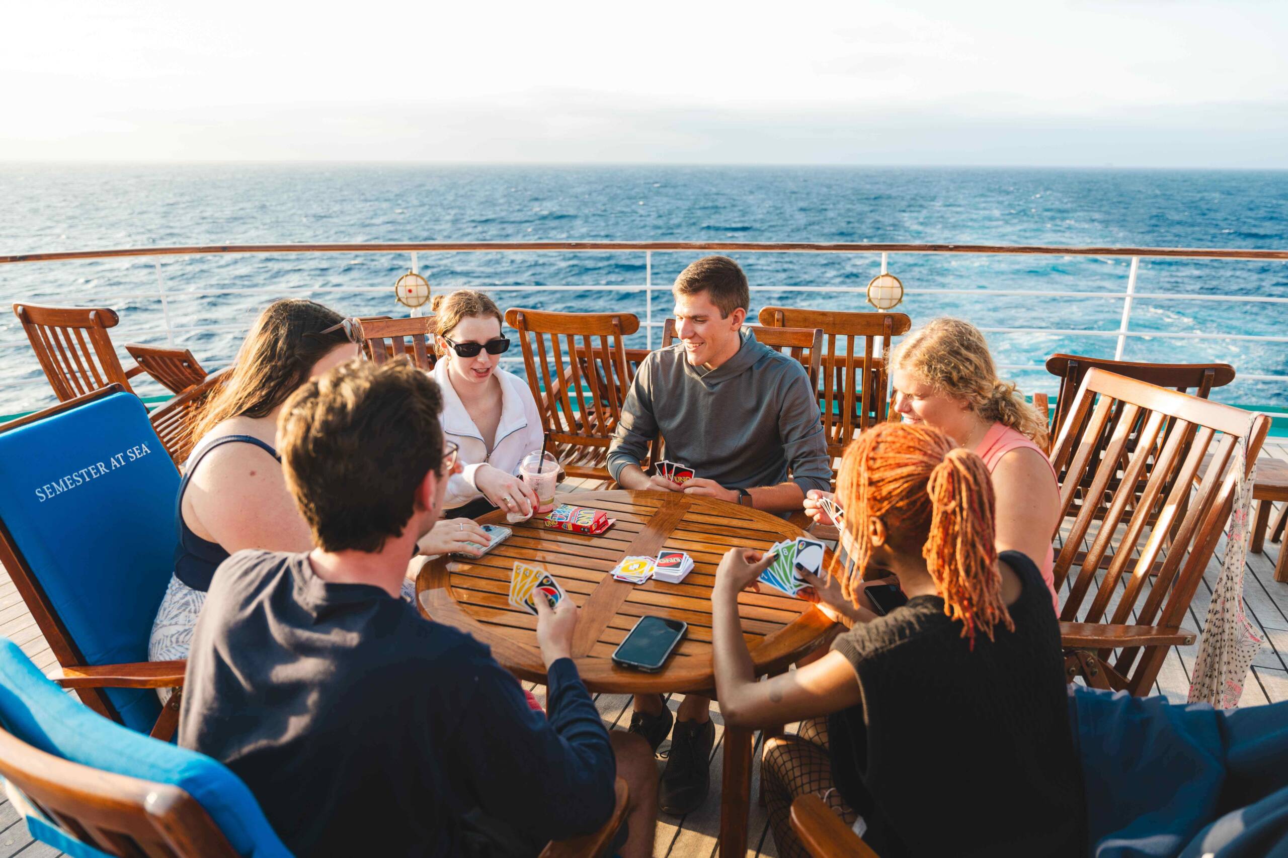 Six people sit around a wooden table on the deck of a ship, playing a card game. A wide blue ocean stretches behind them.