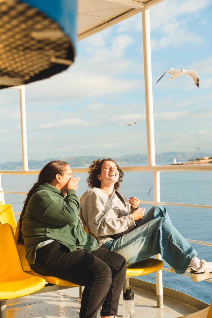 Two people sit side-by-side on yellow chairs on the sunny deck of a ship; both are laughing. Smooth blue water, flying seagulls, and distant green hills are visible beyond the ship’s railing.