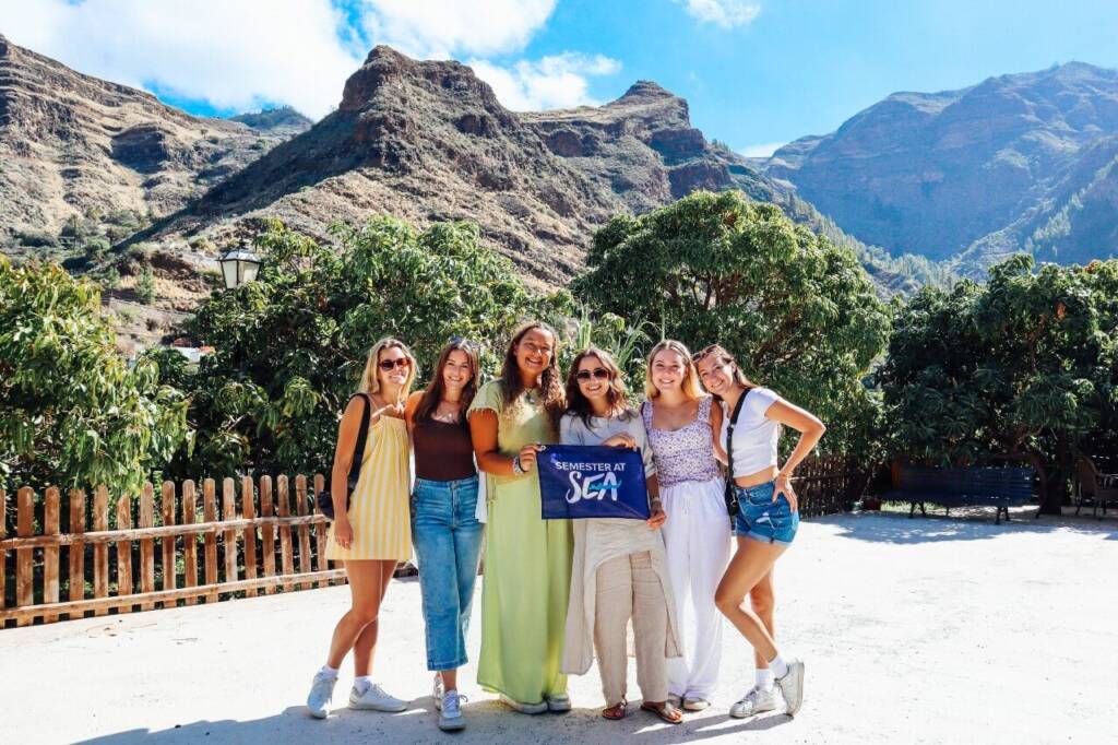 Five young women pose in front of tropical foliage on the edge of a jagged mountain landscape. Two hold a small blue flag that says “Semester at Sea.”