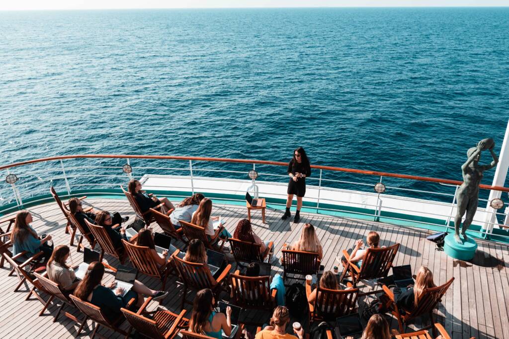 A group of college students sitting on wooden deck chairs on the deck of a ship, attending an outdoor academic lecture. The professor, dressed in black, stands at the front near the ship's railing, with the ocean visible in the background.