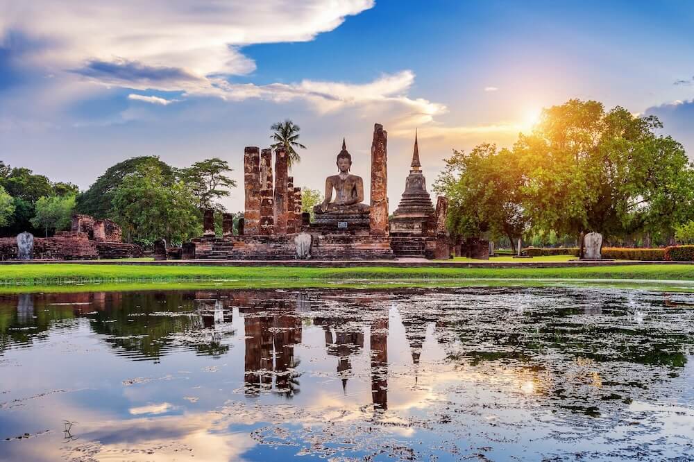 A massive Buddha statue surrounded by stone pillars sits in a grassy park. It is facing a body of water and surrounded by full, green trees.