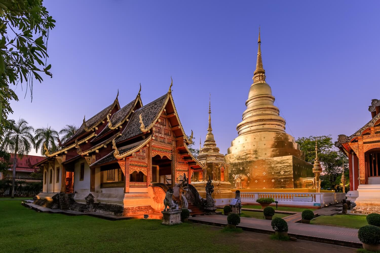 A Buddhist temple with pointed rooflines and ornate gold detailing is illuminated against an indigo twilight sky. It sits next to a large golden structure with a pointed top.