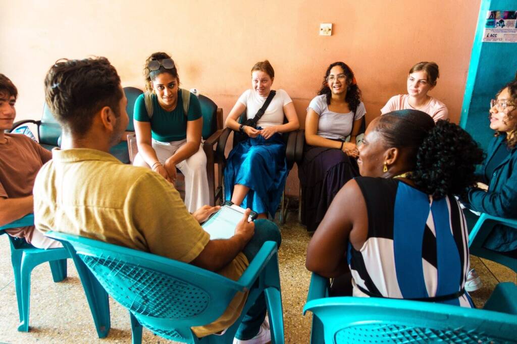 a group of students sit in a circle in teal chairs, active in discussion with one another