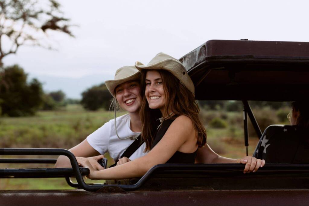 Two young women sit in an open-top vehicle, both wearing safari hats and smiling at the camera.