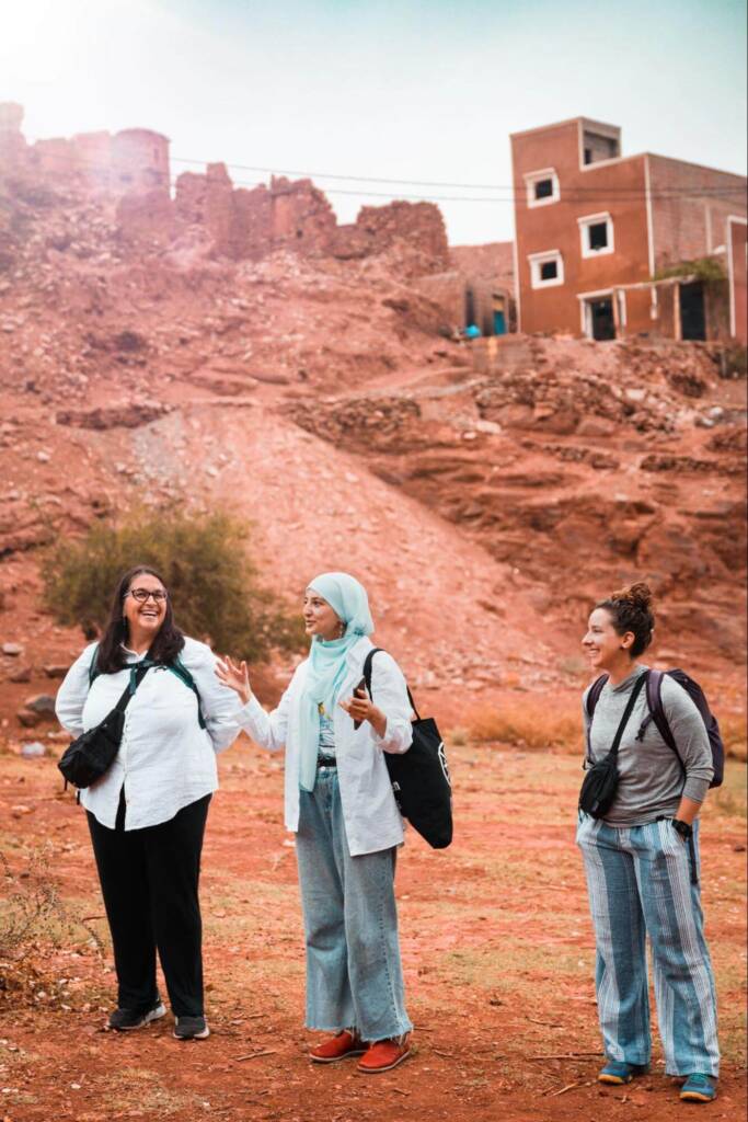 Three women stand side-by-side in front of a red dirt slope that rises behind them. The woman in the center speaks and gestures, while the others listen and laugh.