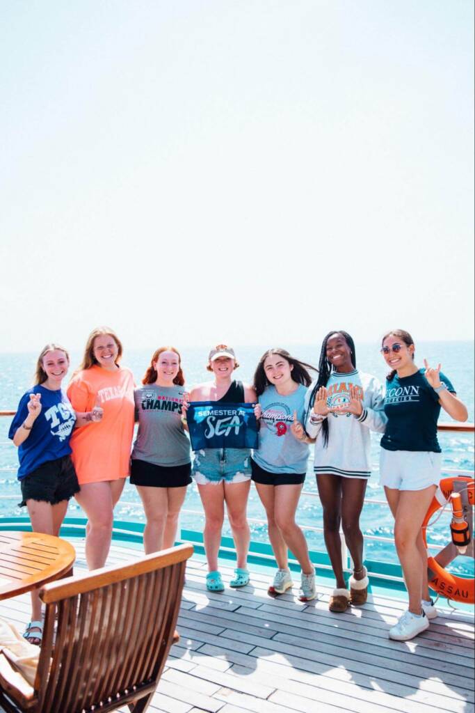 Seven college-aged young people stand in a row on the sunny deck of a ship, each wearing a t-shirt featuring a college logo. The student in the center holds a dark blue flag that says “Semester at Sea.”