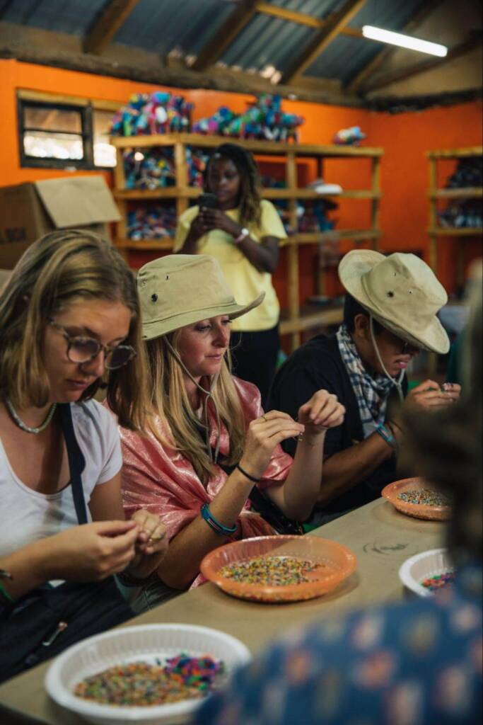 Several young people sit at a table, stringing beads on wire. Plates of colorful beads sit on the table in front of them. 